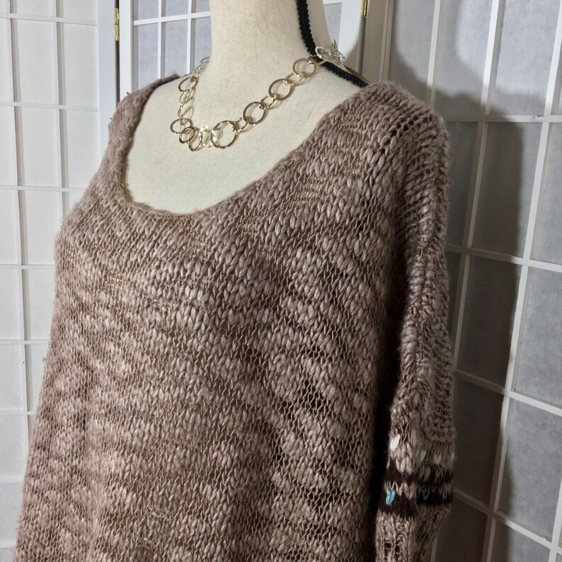 May include: A brown, open-weave sweater with a scoop neck and long sleeves. The sweater is displayed on a mannequin and paired with a silver chain necklace. The sleeves have a patterned design near the cuffs.