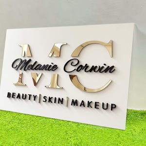 Op de afbeelding: Een wit bord met gouden en zwarte letters. Het bord toont "MC Melanie Corwin" in een scriptlettertype, met "BEAUTY | SKIN | MAKEUP" eronder. De letters "MC" en "IV" zijn goudkleurig, de rest is zwart. Op een groene ondergrond.