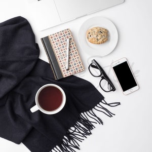 May include: A flat lay of a white laptop, a black scarf, a white cup of tea, a small white plate with a cookie, a black and white notebook with a pen, a pair of black glasses, and a white smartphone.