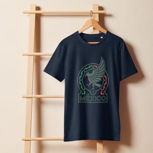 May include: Navy blue t-shirt featuring a gray and green emblem with a stylized eagle and the word "MÉXICO" in gray. The emblem is encircled by green and red lines. The t-shirt is hanging on a wooden hanger.