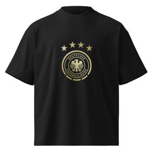 May include: Black t-shirt featuring a gold emblem of the Deutscher Fussball-Bund (DFB), the German Football Association. The emblem includes a golden eagle, stars, and text. The shirt has short sleeves and a crew neck.