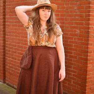 May include: A woman wearing a straw hat, a short-sleeved blouse with a brown and tan floral pattern, and a long, dark brown skirt with a large pocket. The background is a red brick wall.