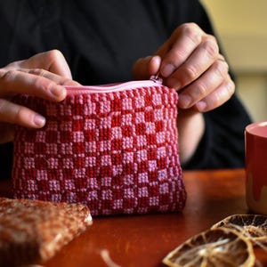 May include: A small, rectangular zipped pouch featuring a woven checkerboard pattern in alternating shades of dark red and light pink. The pouch is constructed from yarn and includes a matching light pink zip closure along the top edge.