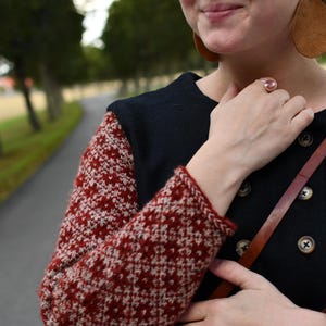 May include: A black long-sleeved sweater featuring a red and white geometric fair isle pattern on the sleeves. The sweater has a double-breasted front closure with dark buttons and a round neckline. The garment is worn with a brown leather strap.