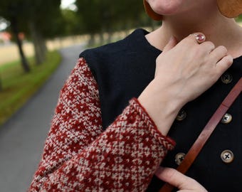 May include: A black long-sleeved sweater featuring a red and white geometric fair isle pattern on the sleeves. The sweater has a double-breasted front closure with dark buttons and a round neckline. The garment is worn with a brown leather strap.