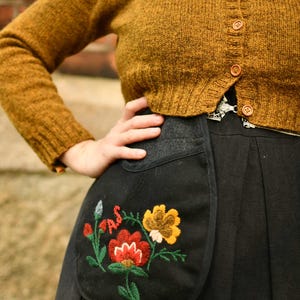 May include: Mustard yellow knit cardigan with wooden buttons. A black skirt with a pocket featuring embroidered flowers in red, yellow, and green. The embroidery includes the letters "A.S."