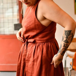 Op de afbeelding: Een roestkleurige linnen jurk met een mouwloos ontwerp en een strikdetail in de taille. De jurk heeft een gerimpelde rok en een eenvoudig, elegant silhouet, geschikt voor casual of semi-formele gelegenheden.