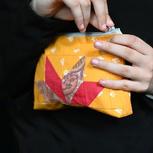 May include: A yellow fabric pouch with a red starburst design and a zipper closure. The pouch has a small pocket on the front and is decorated with white floral patterns. The pouch is held against a black background.