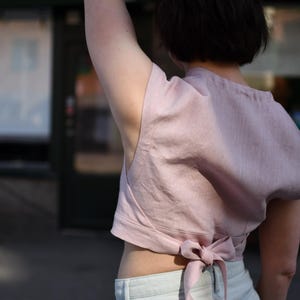 May include: A pale pink sleeveless crop top featuring a high neckline and a tie-back closure at the waist. The garment is constructed from a textured fabric and is paired with light blue denim jeans that have a visible waistband and belt loops.