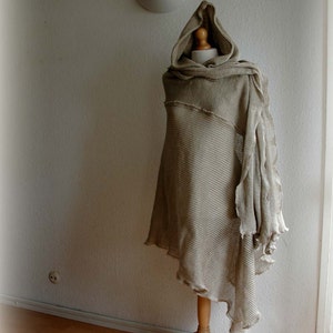 K&ouml;nnte beinhalten: Ein beiger gestrickter Kapuzenponcho mit ausgefransten Kanten, auf einem Holzst&auml;nder vor einer wei&szlig;en Wand ausgestellt.
