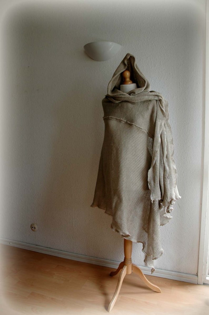 K&ouml;nnte beinhalten: Ein beiger gestrickter Kapuzenponcho mit ausgefransten Kanten, auf einem Holzst&auml;nder vor einer wei&szlig;en Wand ausgestellt.
