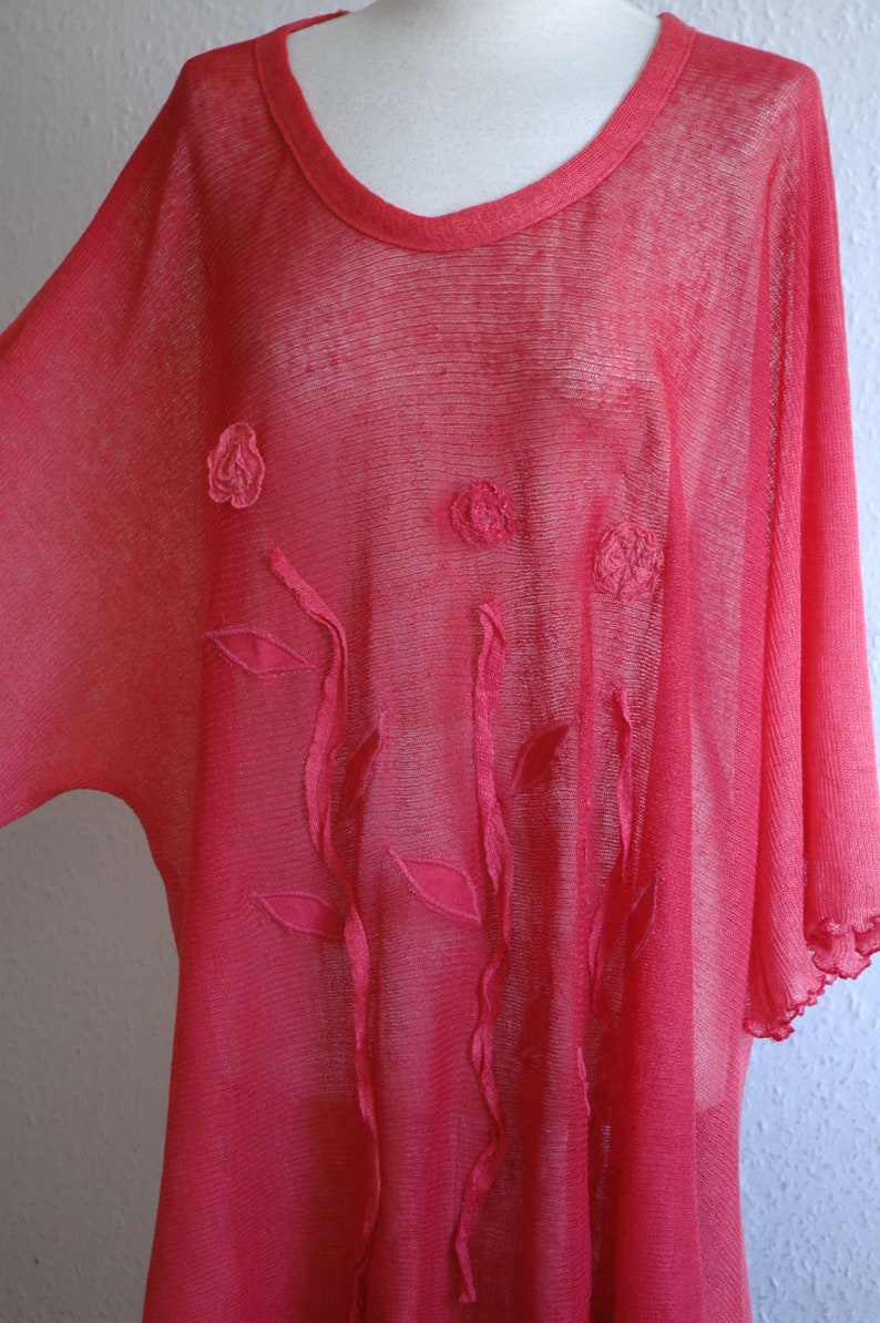 Puede incluir: Una blusa rosa transparente con detalles florales bordados. La blusa tiene un corte holgado y fluido y un escote redondo.