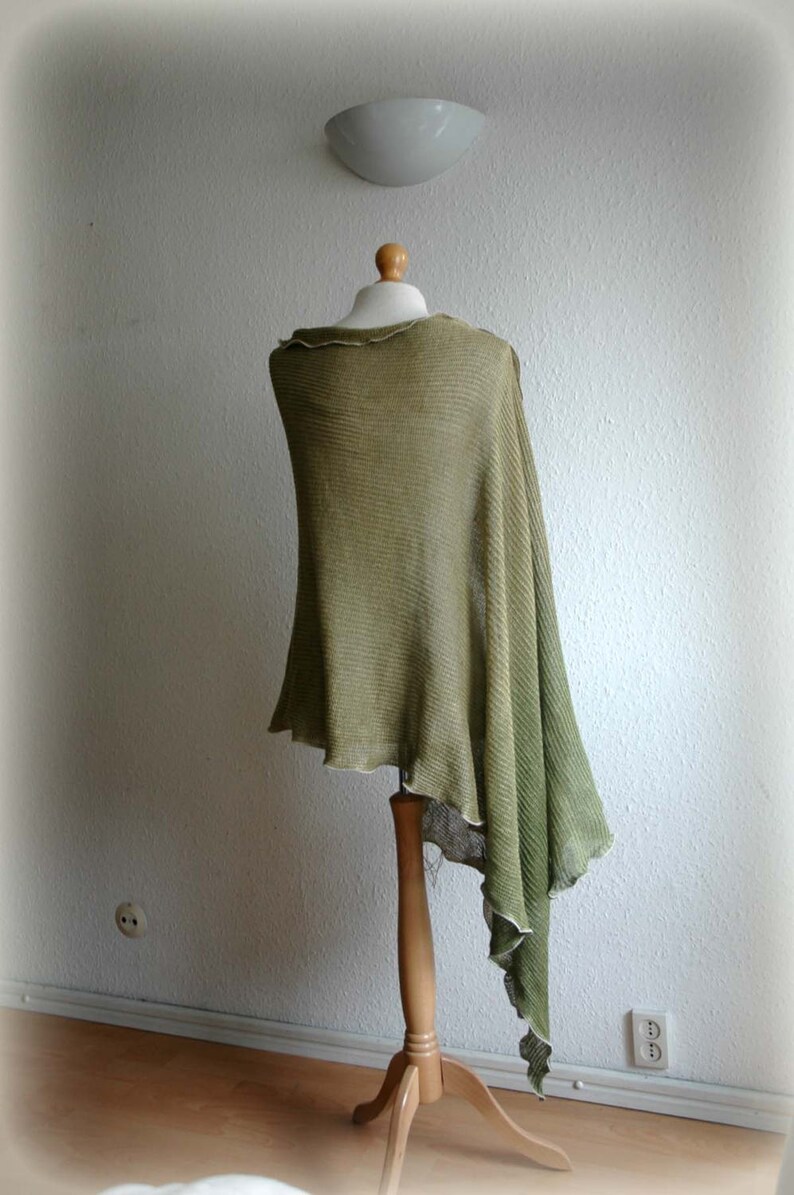 May include: A green knitted poncho draped over a wooden mannequin. The poncho has a loose, flowing fit and a fringed edge.