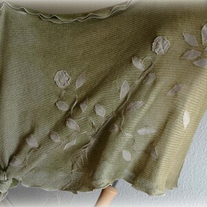 May include: A green knitted shawl with white embroidered floral designs. The shawl has a scalloped edge and is made of a soft, lightweight fabric.