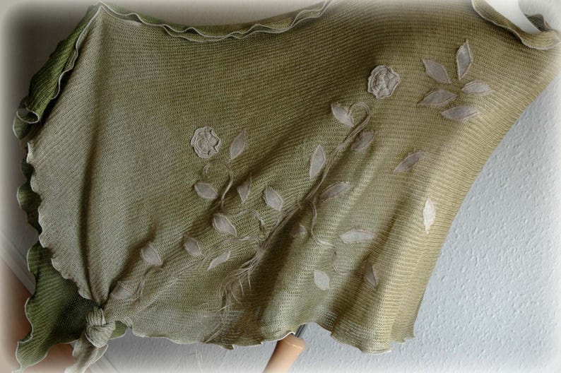 May include: A green knitted shawl with white embroidered floral designs. The shawl has a scalloped edge and is made of a soft, lightweight fabric.
