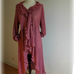 May include: A long, pink cardigan with a ruffled neckline and a decorative flower accent. The cardigan is made of a soft, lightweight fabric and has a flowing, asymmetrical hemline.