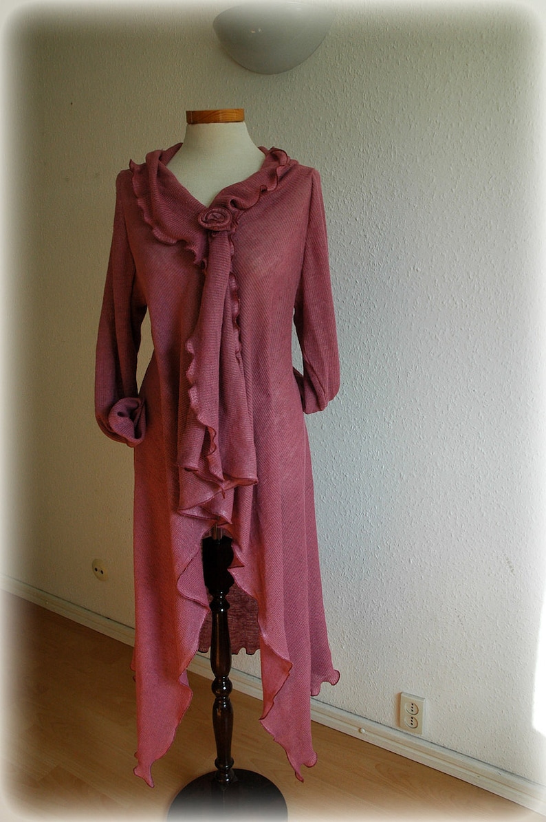 May include: A long, pink cardigan with a ruffled neckline and a decorative flower accent. The cardigan is made of a soft, lightweight fabric and has a flowing, asymmetrical hemline.