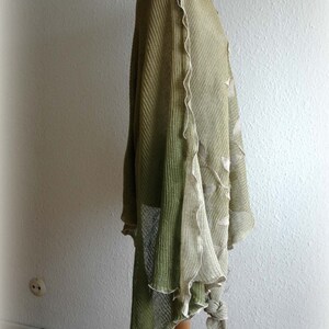 May include: A green and beige knitted shawl with a scalloped edge. The shawl is draped over a mannequin and has a flowing, asymmetrical design.
