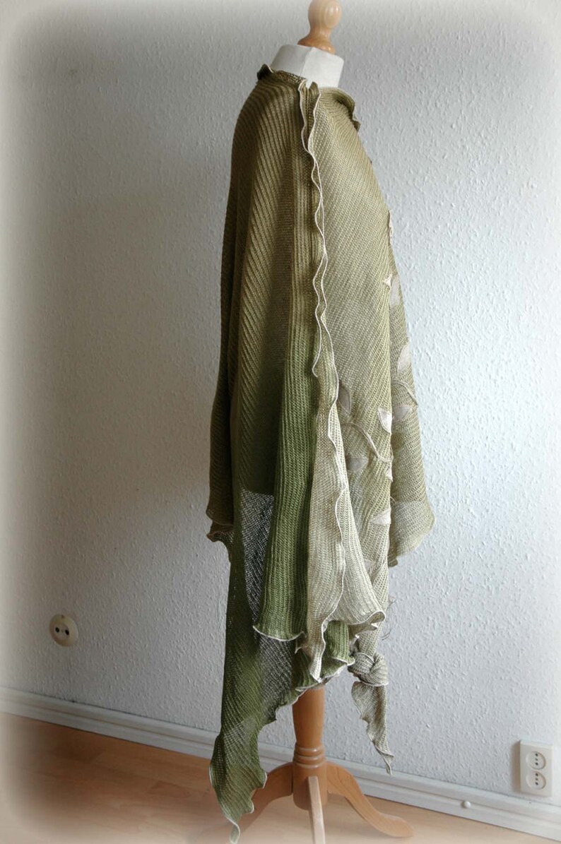 May include: A green and beige knitted shawl with a scalloped edge. The shawl is draped over a mannequin and has a flowing, asymmetrical design.