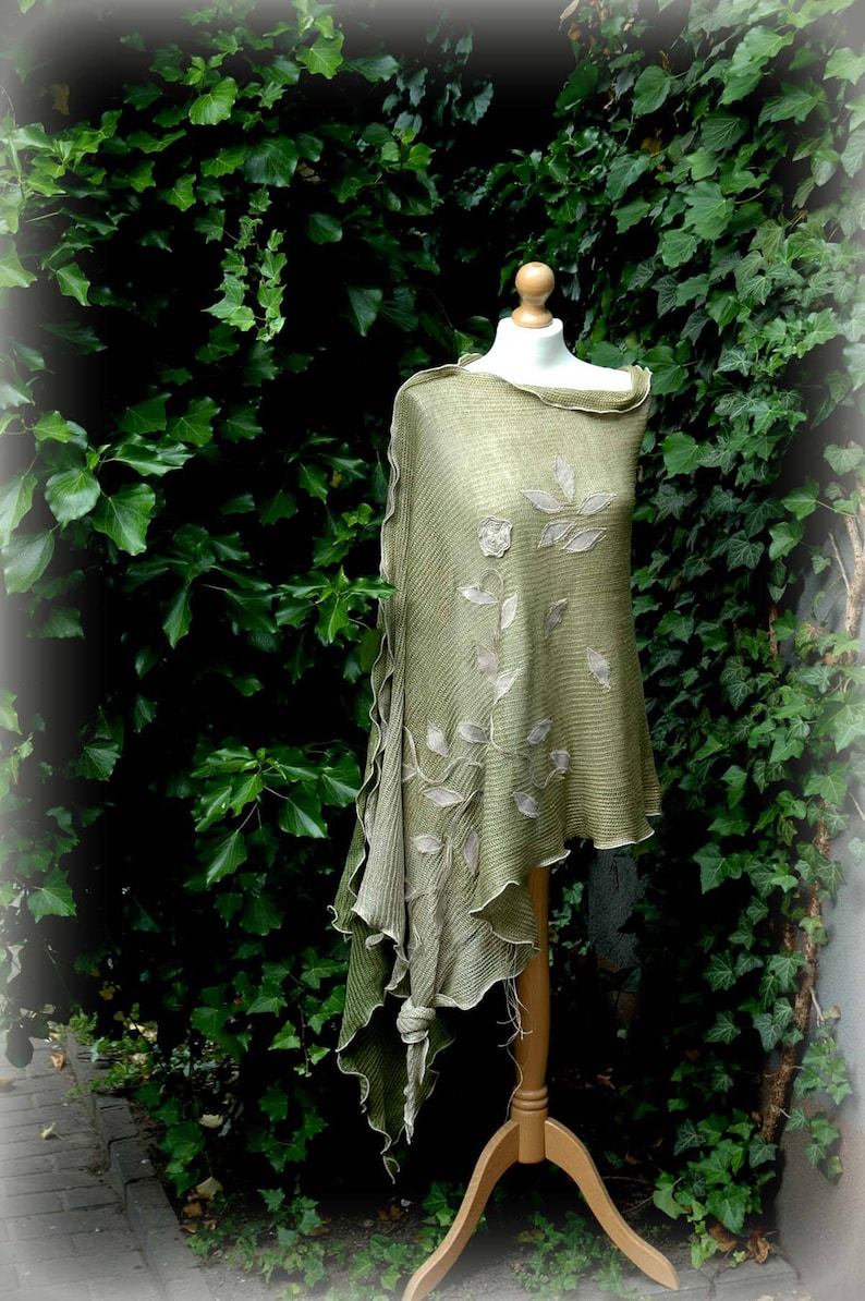 May include: A green knitted shawl with a floral design, displayed on a wooden mannequin against a backdrop of green foliage.