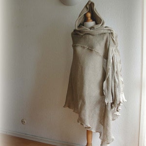 K&ouml;nnte beinhalten: Ein beiger gestrickter Kapuzenponcho mit ausgefransten Kanten, auf einem Holzst&auml;nder vor einer wei&szlig;en Wand ausgestellt.