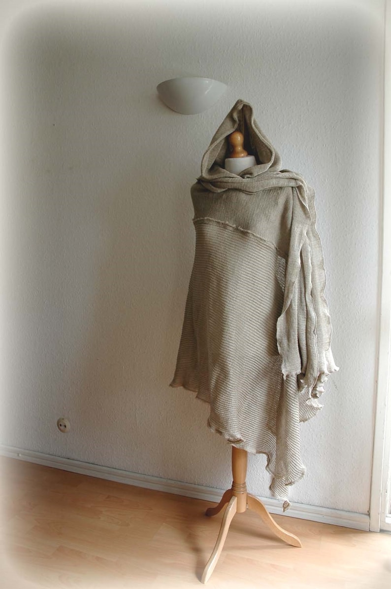 K&ouml;nnte beinhalten: Ein beiger gestrickter Kapuzenponcho mit ausgefransten Kanten, auf einem Holzst&auml;nder vor einer wei&szlig;en Wand ausgestellt.