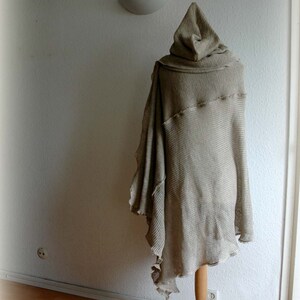 K&ouml;nnte beinhalten: Ein beiger gestrickter Kapuzenponcho, der auf einem Holzst&auml;nder vor einer wei&szlig;en Wand pr&auml;sentiert wird. Der Poncho hat eine lockere, flie&szlig;ende Silhouette und eine lange, spitze Kapuze.