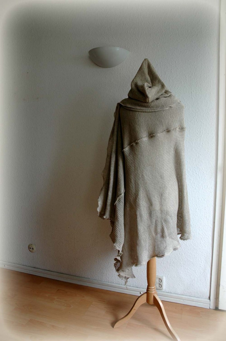 K&ouml;nnte beinhalten: Ein beiger gestrickter Kapuzenponcho, der auf einem Holzst&auml;nder vor einer wei&szlig;en Wand pr&auml;sentiert wird. Der Poncho hat eine lockere, flie&szlig;ende Silhouette und eine lange, spitze Kapuze.