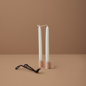 Op de afbeelding: Twee witte kaarsen in een lichtbruine leren kaarsenhouder. Een zwarte metalen kaarsendover ligt op de tafel.