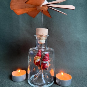 Può includere: Una bottiglia di vetro trasparente con tappo di sughero, che ospita due piccole statuette di gnomi. Una girandola di legno è posta sopra la bottiglia. Due candele a lumino accese affiancano la bottiglia, creando una luce calda.