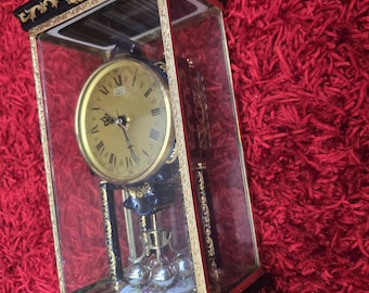 Op de afbeelding: Een rechthoekige schoorsteenklok met een glazen behuizing, voorzien van een goudkleurige wijzerplaat met zwarte Romeinse cijfers en sierlijke gouden filigraanaccenten op een donkerblauwe voet. De klok rust op vier kleine voetjes en heeft een gouden topornament.