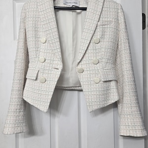 May include: A white tweed blazer with a subtle multi-colored grid pattern, featuring a double-breasted front with six round cream-colored buttons, two flap pockets, and a white interior lining. The inner label reads Veronica Beard.