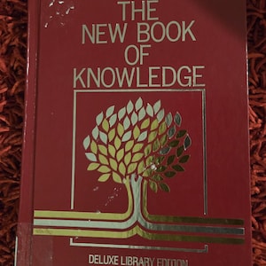May include: A red hardback book titled The New Book of Knowledge Deluxe Library Edition. The cover features gold and silver foil text and a stylised tree graphic with metallic leaves enclosed in a rectangular frame with horizontal lines.