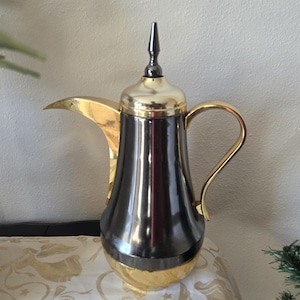 May include: A traditional coffee pot with a gold spout, handle, and base, and a dark, metallic body. The pot has a tall, pointed lid and sits on a patterned surface.