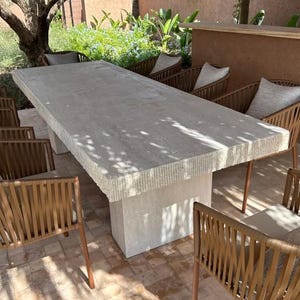 Peut inclure: Une grande table rectangulaire en pierre avec un bord texturé, soutenue par une base solide. Plusieurs chaises à dossier tressé avec coussins entourent la table, créant un ensemble de salle à manger extérieur. La table mesure environ 3 mètres de long.