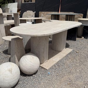Peut inclure: Ensemble de meubles en pierre, comprenant des tables et des sphères décoratives. Les tables, de couleur beige clair, reposent sur des bases cylindriques ou rectangulaires. Les sphères, également beige clair, présentent une surface texturée.
