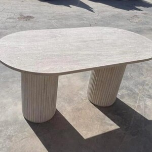 Peut inclure: Une table ovale de couleur claire avec un plateau en pierre et deux pieds cylindriques cannelés. La table est faite d'une pierre de couleur claire. Ses dimensions sont d'environ 152 cm de long et 76 cm de large. Pour l'intérieur ou l'extérieur.
