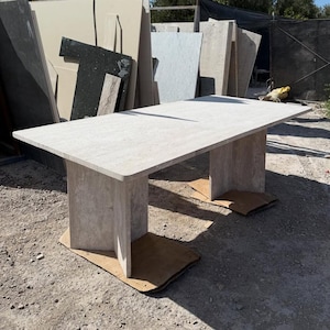 Peut inclure: Une table rectangulaire en pierre de couleur claire avec une surface lisse. La table a deux pieds rectangulaires épais. La table est à l'extérieur, avec d'autres dalles de pierre en arrière-plan.