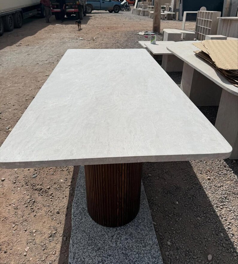May include: A rectangular stone table with a light grey top and a dark brown, cylindrical base. The base has a vertical, ribbed design. The table is outdoors on a gravel surface.