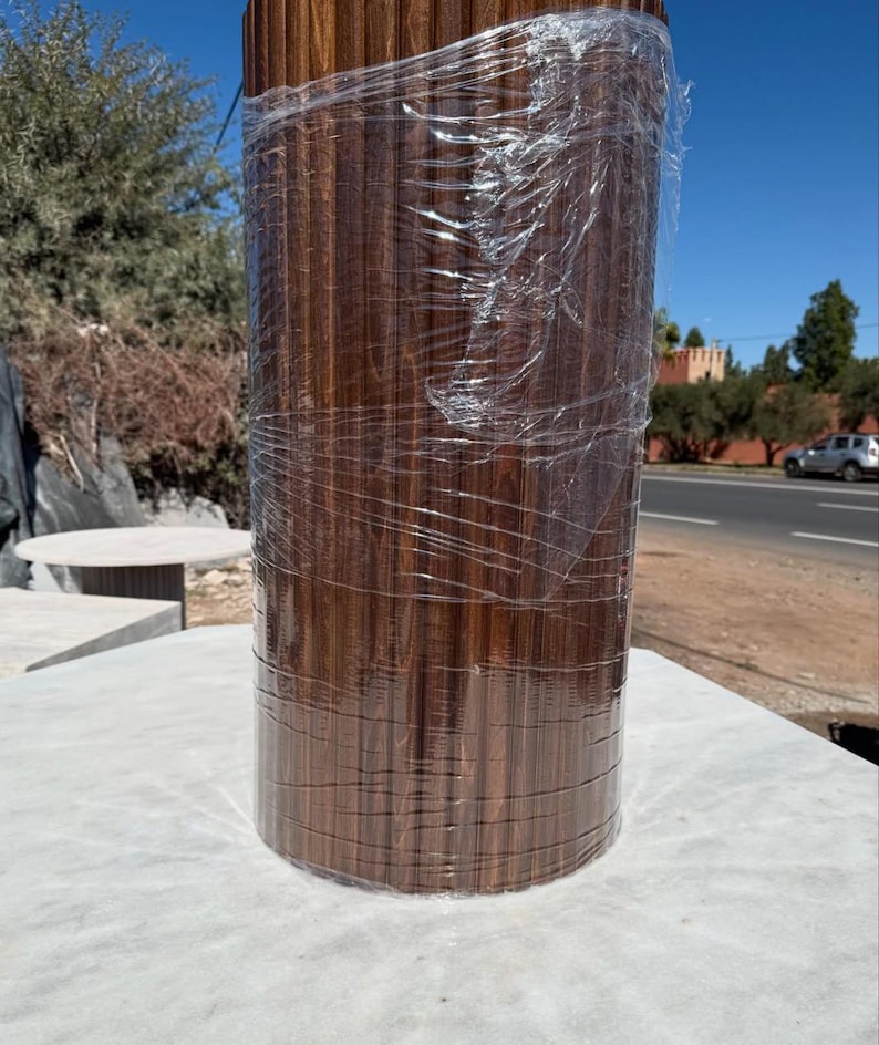 May include: A cylindrical wooden product, wrapped in clear plastic, stands on a white surface. The wood has a rich, dark brown colour with vertical grain patterns. The product appears to be new, possibly for construction or interior design.