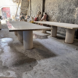 May include: Two large, light-colored rectangular tables with thick, cylindrical legs. The tables are in a workshop setting with a concrete floor and walls. The tables appear to be made of wood or a similar material.