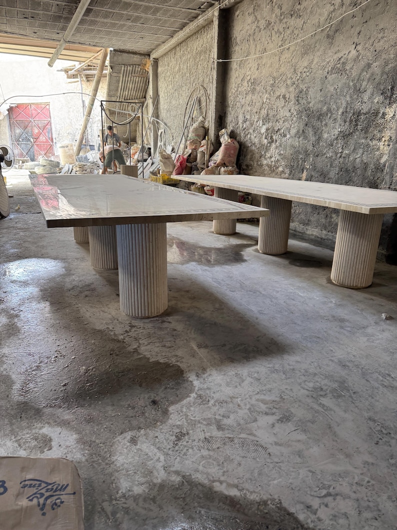 May include: Two large, light-colored rectangular tables with thick, cylindrical legs. The tables are in a workshop setting with a concrete floor and walls. The tables appear to be made of wood or a similar material.
