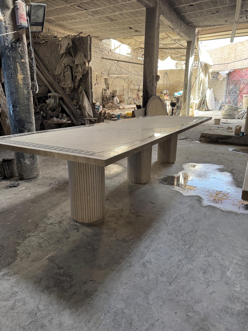 May include: A large rectangular marble table with a decorative border. The table is supported by three cylindrical, fluted legs. The table appears to be in a workshop or studio setting.