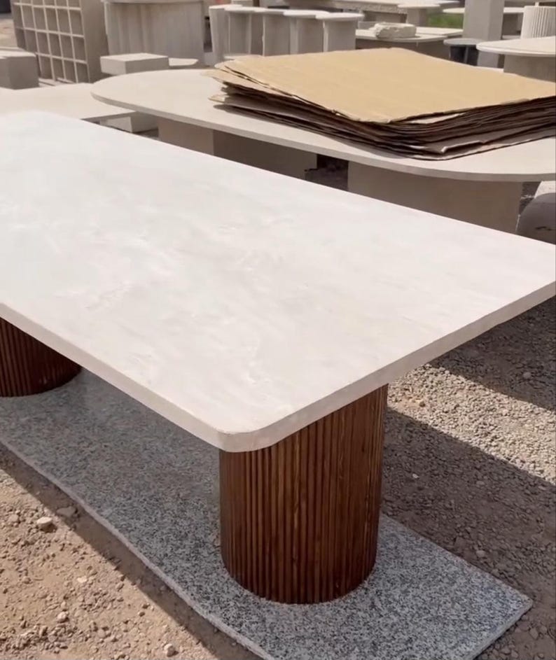 May include: A rectangular table with a light-coloured stone top and two cylindrical, dark brown wooden legs. The table is set on a grey surface, with other tables visible in the background. The table's dimensions are not visible.