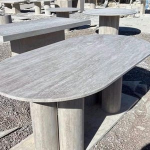 Peut inclure: Plusieurs tables en pierre de différentes formes et tailles sont présentées à l'extérieur. Les tables ont des dessus et des bases gris clair. Une table a un plateau ovale soutenu par des pieds cylindriques.