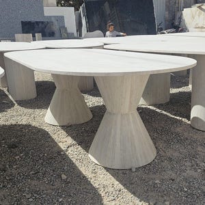Peut inclure: Plusieurs tables en pierre gris clair avec des bases en forme de sablier et des plateaux arrondis. Les tables sont à l'extérieur sur une surface de gravier. Les tables sont en pierre et de couleur neutre.
