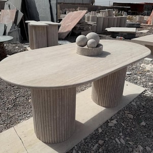 Peut inclure: Une table ovale en pierre beige clair avec deux pieds cylindriques à rainures verticales. Un petit bol rond sur la table contient trois sphères en pierre. La table est à l'extérieur, avec d'autres pièces en pierre en arrière-plan. La table mesure environ 200 cm de long.