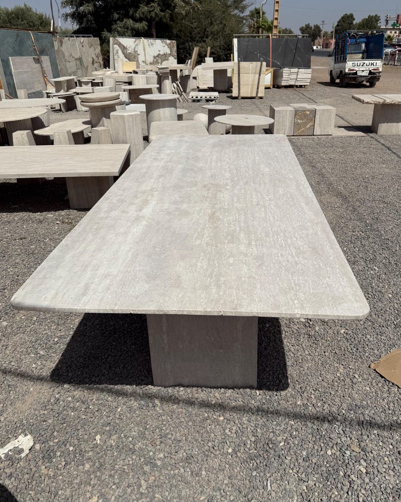 Pu&ograve; includere: Un grande tavolo rettangolare in pietra di colore grigio chiaro. Il tavolo ha una base rettangolare solida. Altri tavoli e panche in pietra sono visibili sullo sfondo, suggerendo mobili da esterno.