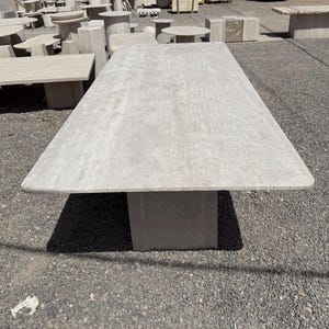 Pu&ograve; includere: Un grande tavolo rettangolare in pietra grigio chiaro con una base rettangolare spessa. Il tavolo &egrave; all'aperto, con altri tavoli e strutture in pietra visibili sullo sfondo. La superficie ha un aspetto strutturato.