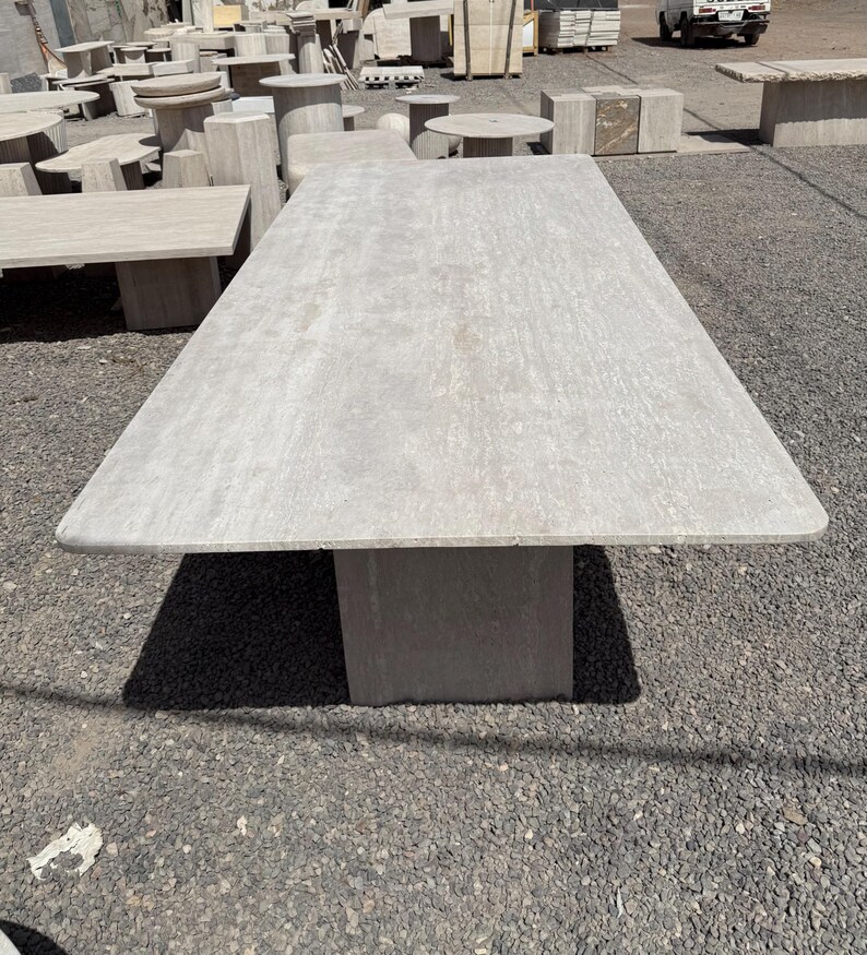 Pu&ograve; includere: Un grande tavolo rettangolare in pietra grigio chiaro con una base rettangolare spessa. Il tavolo &egrave; all'aperto, con altri tavoli e strutture in pietra visibili sullo sfondo. La superficie ha un aspetto strutturato.